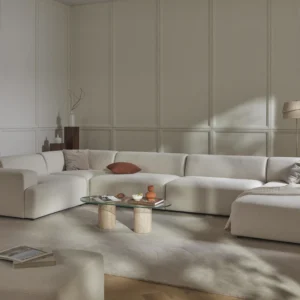 Large L-shaped MELVA lounge sofa in light beige fabric with rounded edges, wide armrests, and deep seat, shown frontally with the corner section on the left.