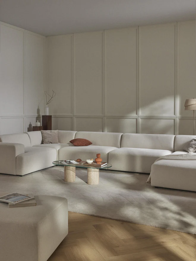 Large L-shaped MELVA lounge sofa in light beige fabric with rounded edges, wide armrests, and deep seat, shown frontally with the corner section on the left.